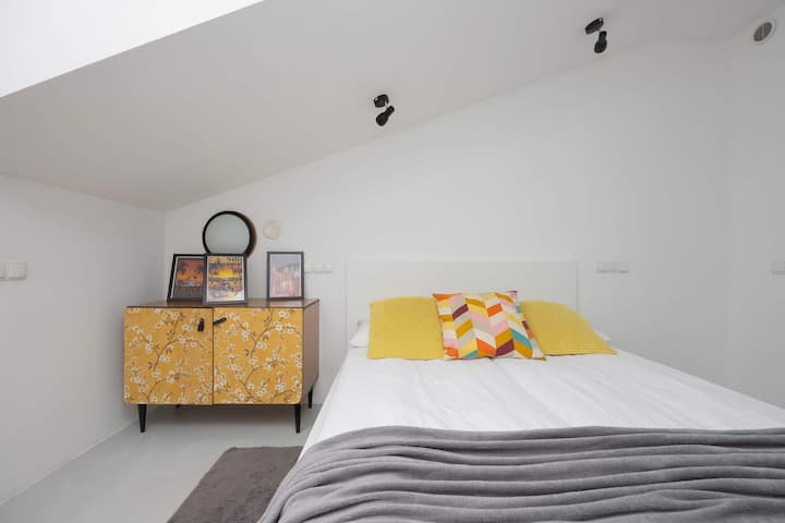 Un dormitorio luminoso con una cama doble, techo inclinado y un pequeño armario naranja para mayor color.