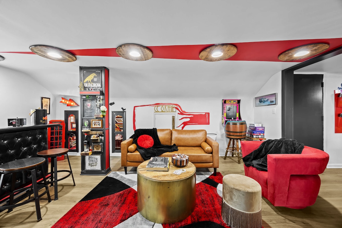 The living area features a comfortable leather couch and a striking red accent chair, complemented by a vibrant area rug. A round coffee table sits at the center, surrounded by shelves displaying various games and decor items. Recessed lighting enhances the modern design.