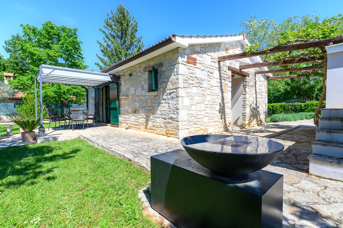 A quaint stone building is highlighted under clear blue skies, surrounded by lush greenery. A modern water feature adds a sleek contrast to the natural setting. Outdoor seating is visible, providing a comfortable space to enjoy the garden vistas.