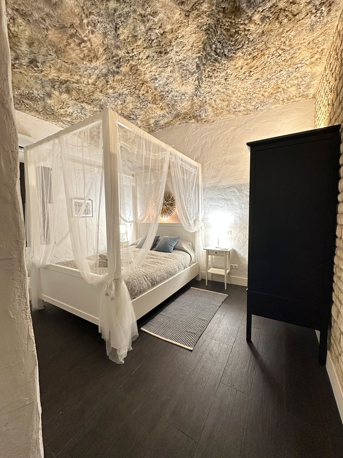 A cozy bedroom features a four-poster bed draped in light fabric, enhancing the serene atmosphere. A simple bedside table with a lamp is positioned next to the bed, and a tall black wardrobe is seen against the textured wall, complemented by a natural stone ceiling.