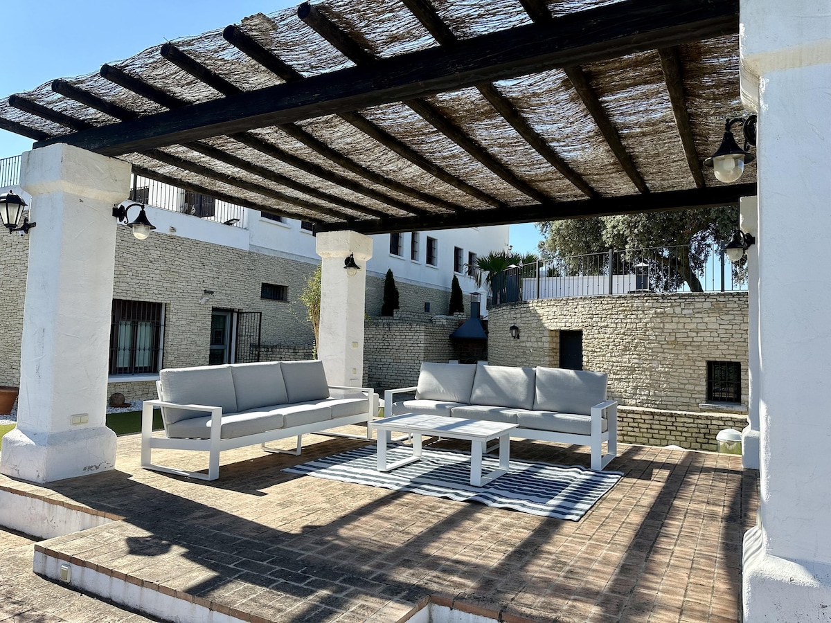 An outdoor shaded seating area is highlighted, featuring comfortable grey sofas arranged around a rectangular table. The space is surrounded by a rustic stone wall and elevated greenery, providing a peaceful atmosphere for relaxation.