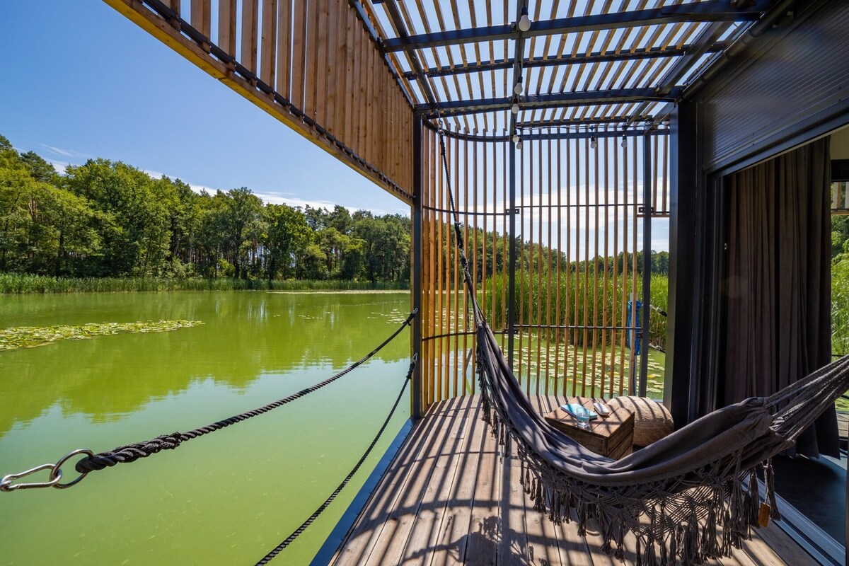 A spacious terrace extends over the water, featuring a hammock and wooden seating for relaxation. Natural light streams through the slatted pergola, creating a play of light and shadow. Lush greenery surrounds the serene lake, enhancing the tranquil atmosphere.