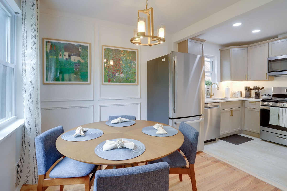 A modern kitchen and dining area is depicted, featuring a round wooden table set with blue placemats and neatly folded napkins. Surrounding the table are four blue upholstered chairs. Wall art in vibrant colors hangs above the table, while stainless steel appliances and cabinetry complement the space.