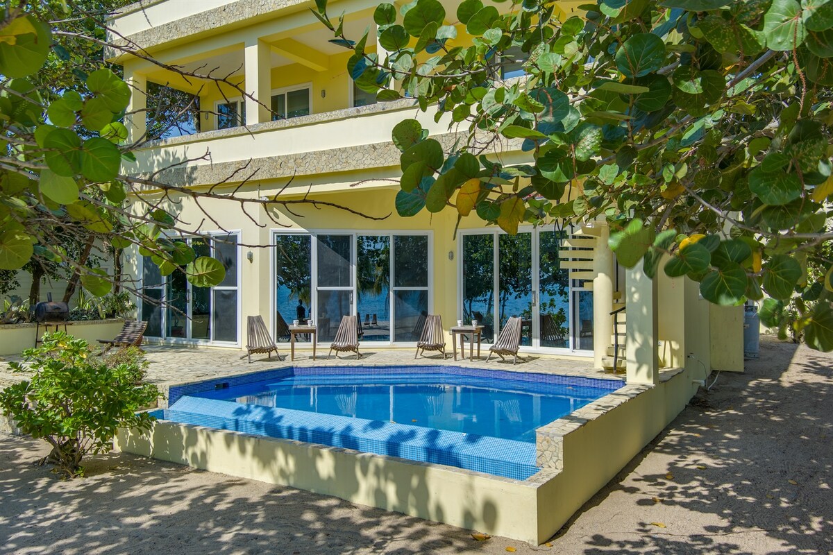 The villa’s exterior features a private above-ground pool bordered by a tiled deck, surrounded by tropical foliage. The bright yellow structure has large glass doors offering views of the pool and beach beyond, creating a seamless connection with the outdoor area.