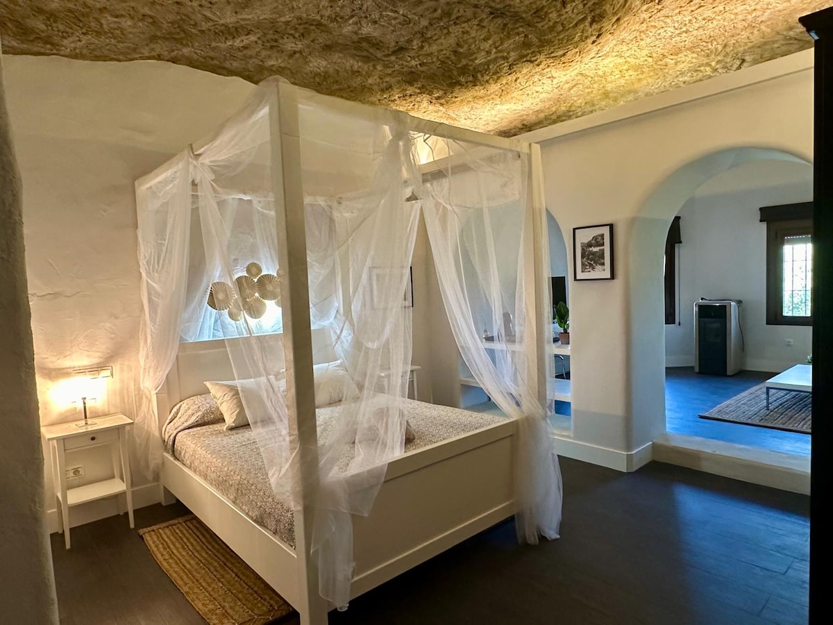 A spacious bedroom features a four-poster bed adorned with sheer white drapery. Soft lighting from a bedside lamp illuminates the room, highlighting the textured ceiling and natural stone walls. An arched doorway leads to another area, enhancing the openness of the space.
