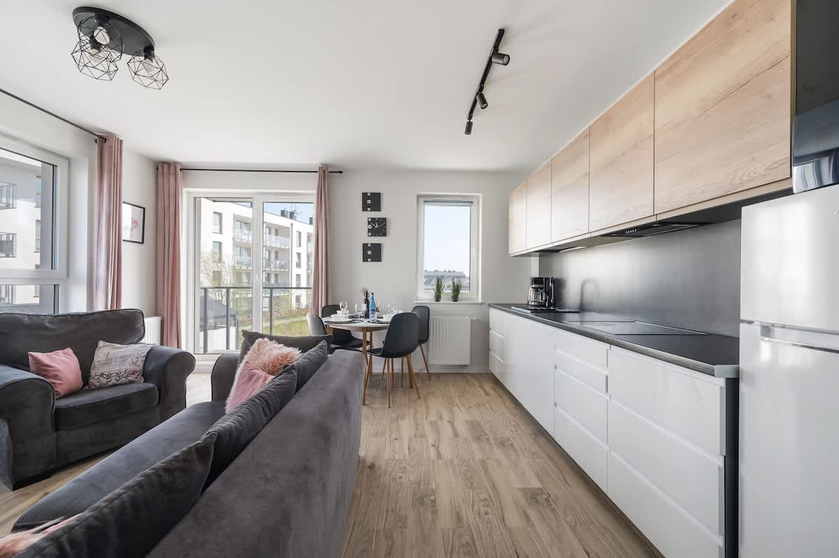 A modern kitchen area is visible, featuring sleek cabinetry and a minimalist design. A dining table with chairs is positioned next to a large window, allowing natural light to fill the space. The adjacent living area is furnished with a comfortable sofa and decorative cushions.