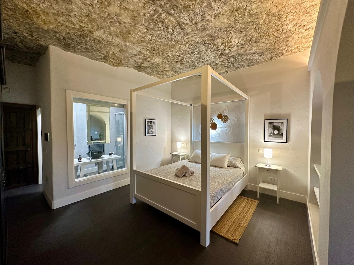 A spacious bedroom features a four-poster bed dressed in soft linens, complemented by bedside tables with lamps. A large mirror reflects the room's minimalist design, with textured ceilings adding depth. Natural light illuminates the space, enhancing the calm and inviting environment.
