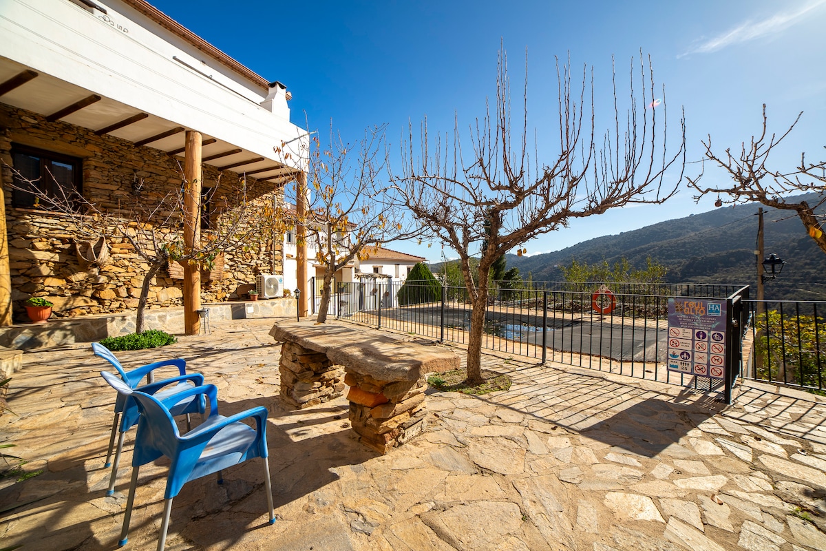 An outdoor terrace features a stone table surrounded by two blue chairs. Leafless trees are seen on either side, with a backdrop of mountains and clear skies, creating an inviting space for relaxation.