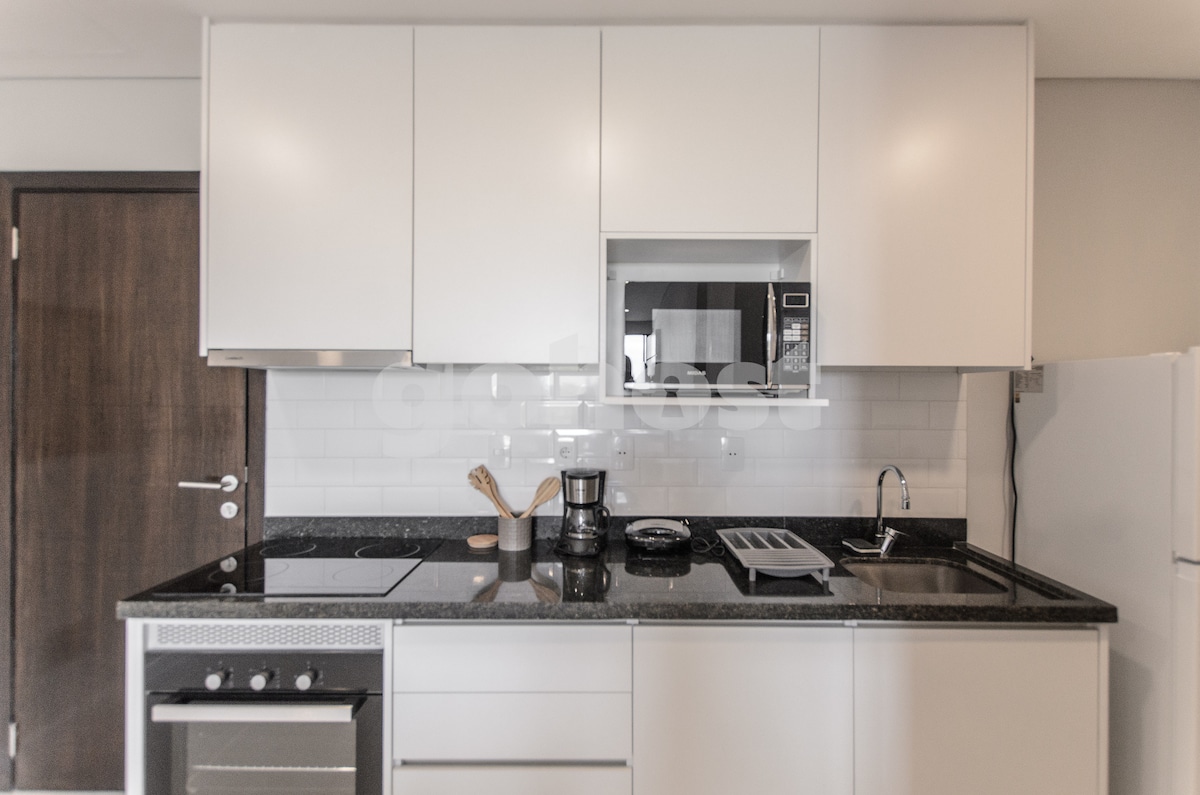 A modern kitchen area features sleek white cabinetry and a contrasting dark countertop. An electric stove, microwave, and coffee maker are visible, alongside essential kitchenware. A small set of utensils is neatly arranged, contributing to a clean and organized cooking space.