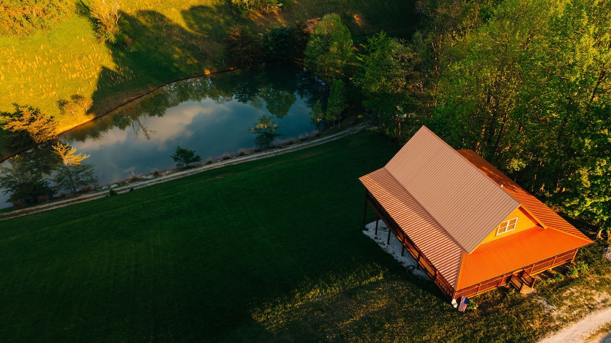 An aerial view of a rustic cabin is showcased beside a serene private pond, surrounded by verdant grass and trees. A gently sloped hillside is visible in the background, enhancing the tranquil setting of this peaceful retreat.