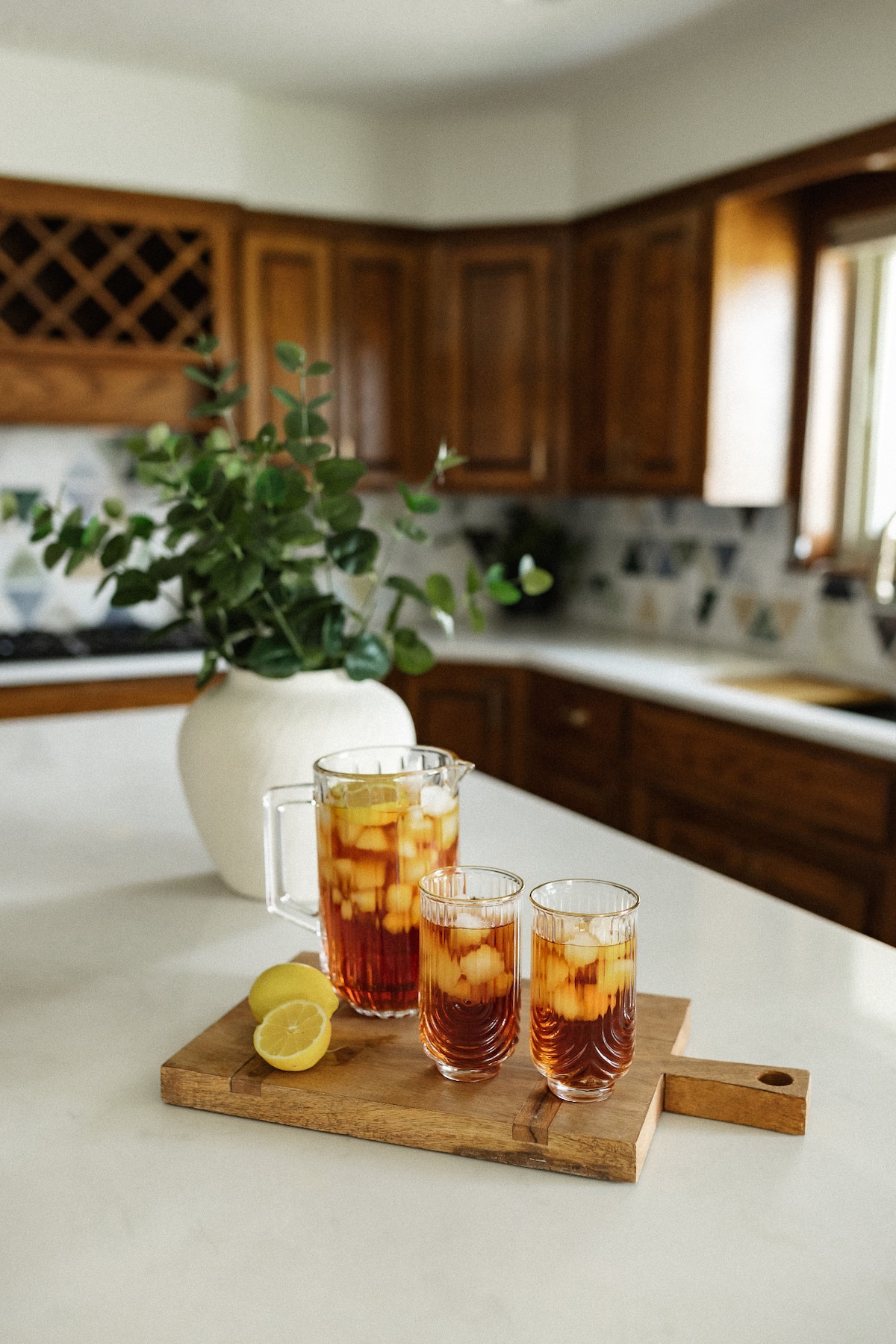 A wooden serving board holds a pitcher of iced tea with lemon slices and two tall glasses filled with the drink. A sprig of greenery in a round vase sits nearby on a light marble countertop, complemented by warm wooden cabinetry in the background.