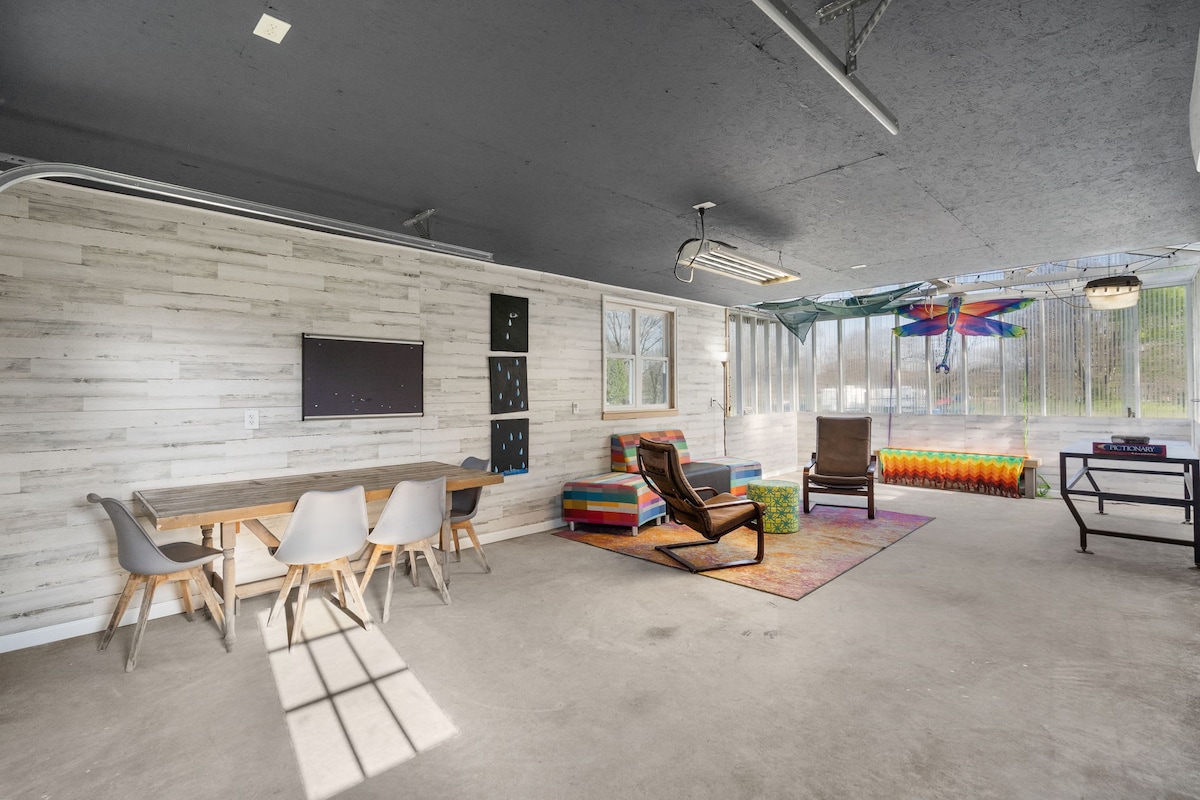 An open garage space features a large window wall allowing natural light to fill the room. A dining table with chairs sits alongside a colorful seating area with cushions. Brightly patterned rugs add a playful touch to the concrete floor.
