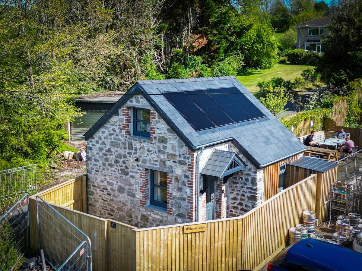 A charming stone cottage is embraced by a wooden fence that ensures privacy. Solar panels are mounted on the sloped roof, reflecting sustainability. The surrounding area is lush with greenery, with outdoor seating nearby, inviting relaxation amidst nature.