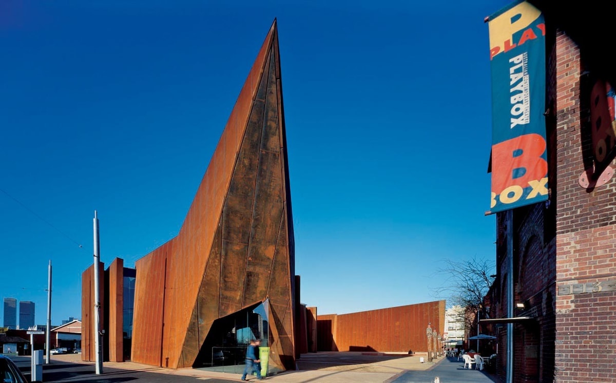 A modern architectural structure features a striking angular facade composed of rusted metal. The building's sharp lines create a dynamic visual contrast against a clear blue sky, while a colorful sign is visible nearby, enhancing the lively ambiance of the surrounding area.
