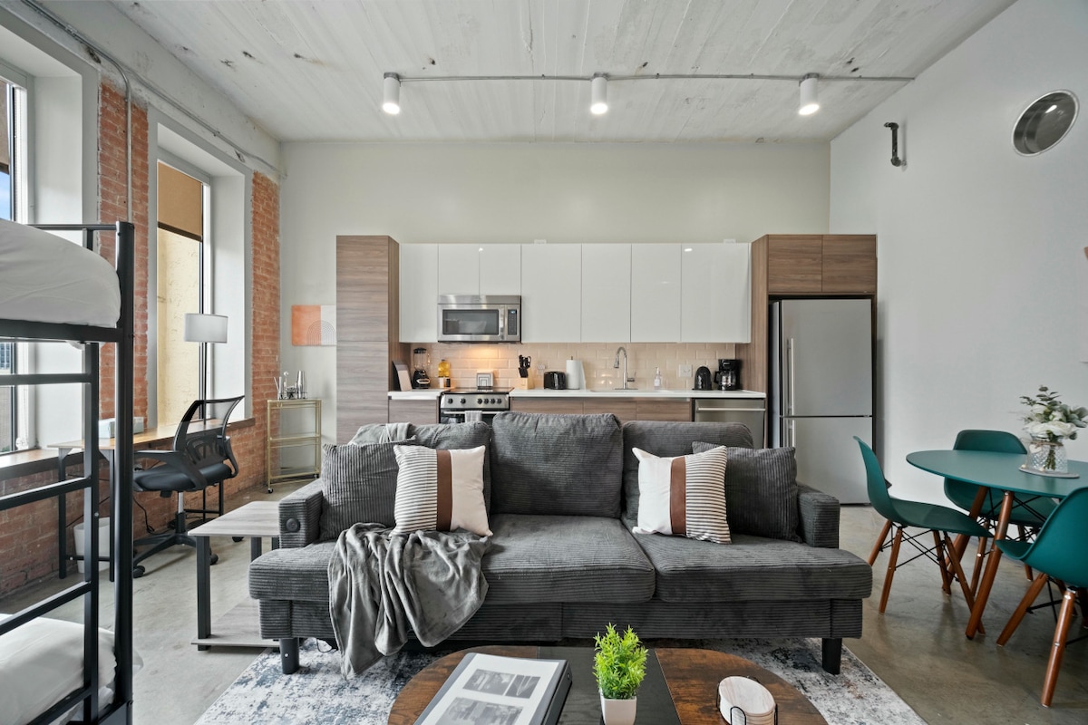 A spacious living area features a comfortable sofa adorned with decorative pillows. The open kitchen contains modern appliances, including a refrigerator and microwave. A dining table is situated nearby, surrounded by teal chairs. Natural light filters through large windows, enhancing the inviting space.