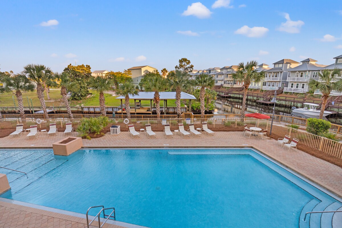 A clear blue pool is framed by sun loungers, offering a relaxing space for guests. Lush palm trees surround the area, with a boardwalk visible in the background, connecting to a calm waterway. Soft clouds float above a bright sky.