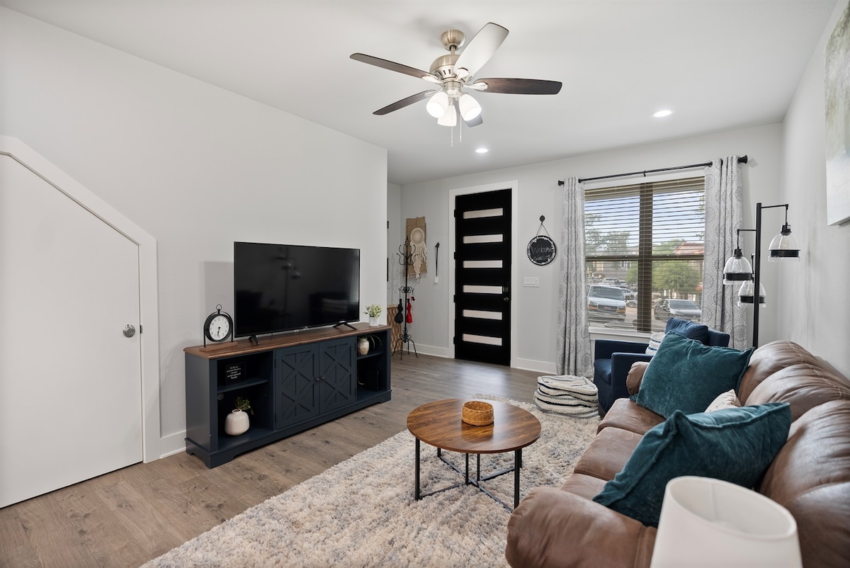 An open living area is showcased, featuring a comfortable brown sofa with teal pillows. A round coffee table sits in front of the sofa, while a flat-screen television is mounted on a dark console. Large windows provide ample natural light, framed by sheer curtains.