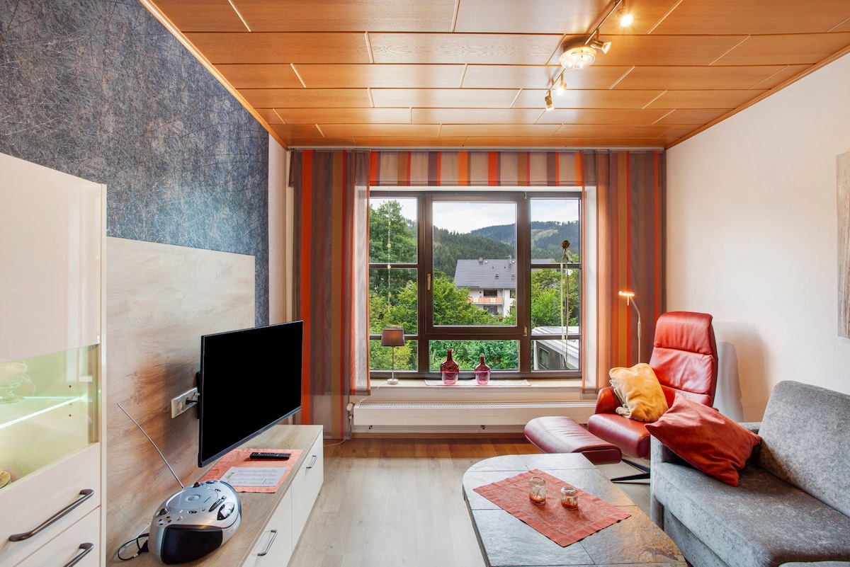 A living room is featured with a large window providing views of greenery outside. A comfortable grey sofa and a red lounge chair are positioned around a coffee table. A television sits on a white cabinet, and bright wall art adds color to the space.