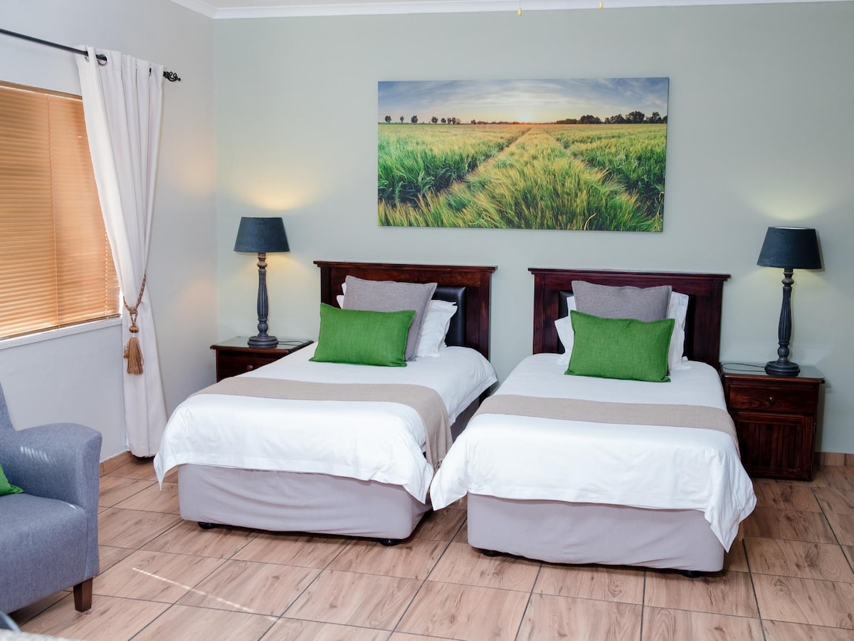 The spacious sleeping area features two 3/4 beds with light bedding and green accent pillows. A landscape painting hangs above the beds, complementing the muted walls. Side tables with lamps are positioned beside each bed, and a comfortable seating area is visible in the corner.