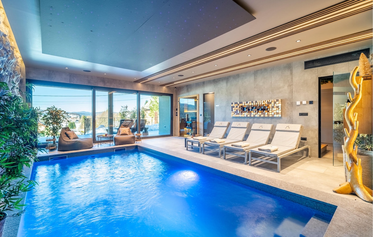 An indoor pool area features a shimmering blue pool surrounded by comfortable lounge chairs. Large glass doors provide natural light and an unobstructed view of the outdoors. Walls are adorned with modern art, and lush green plants add a touch of nature.