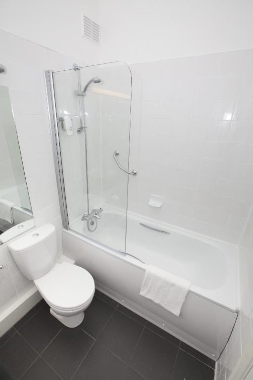 A clean and modern bathroom is highlighted by a glass shower enclosure above a white bathtub. A large mirror is positioned above the sink area, and neutral tiles cover the floor, contributing to a bright and spacious appearance.