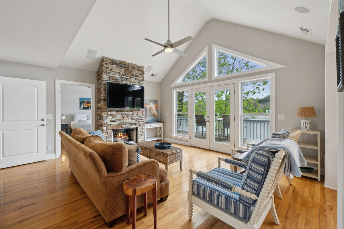 A bright living area features large windows overlooking the river, allowing natural light to fill the space. A stone fireplace is centered on one wall, complemented by a smart TV above it. Comfortable seating arrangements, including a cozy sofa and stylish chairs, complete the inviting atmosphere.