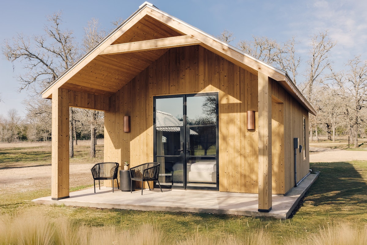 A tranquil cabin exterior features a light wood facade with a sloped roof. Two black chairs and a small table are positioned on a concrete patio, offering a welcoming outdoor space. Large glass doors provide views of the lush grasslands and allow natural light to enter.
