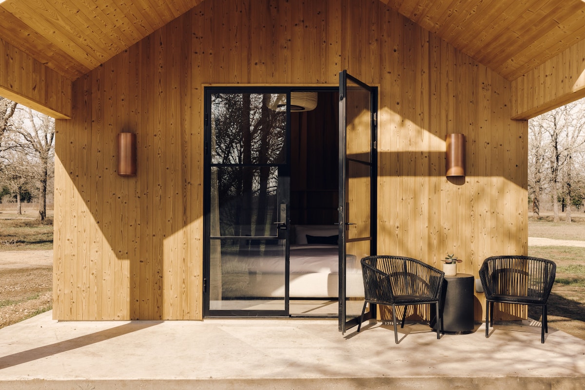 An inviting entrance to a Post Oak Cabin is framed by warm wood paneling. A pair of black wicker chairs are positioned on a concrete patio, complemented by a small side table. Large glass doors open to the interior, allowing natural light to flow in.