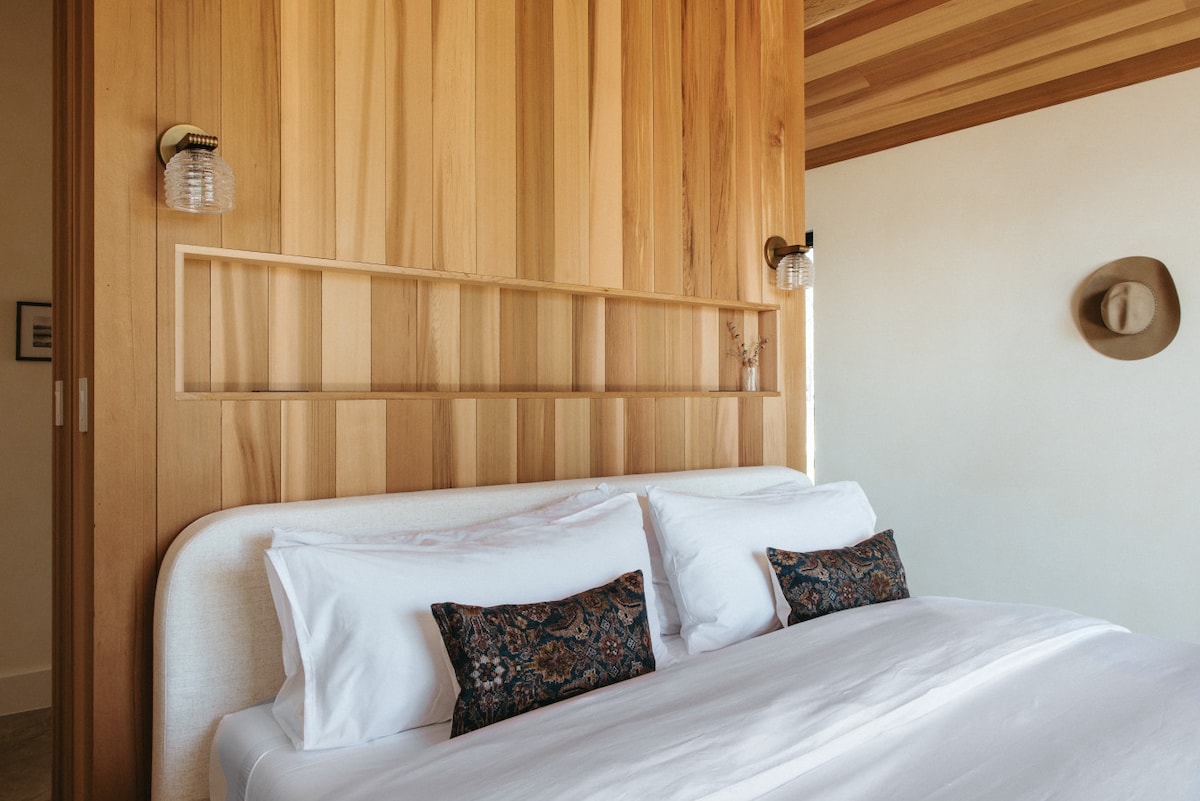 A warm-toned wooden feature wall is set behind a spacious bed, which is dressed in crisp white linens. Two decorative pillows add subtle color. Wall-mounted lights are positioned on either side, and a hat decorates the wall above.