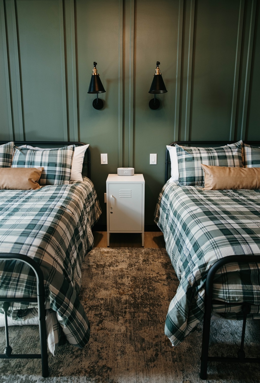 A shared bedroom features two beds with plaid bedding, accompanied by wall-mounted sconces above each bed. A nightstand is placed between the beds, while the room is accented by a dark green wall and a neutral-toned area rug.