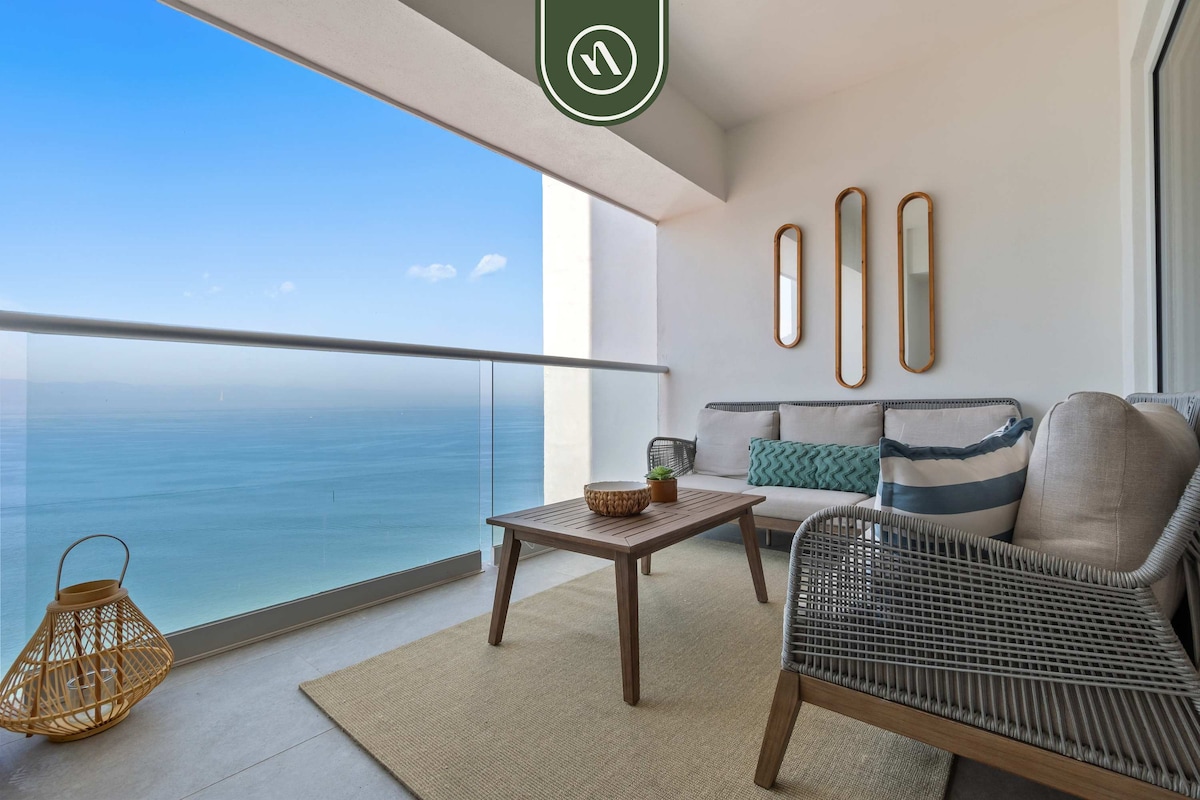 A private terrace features comfortable outdoor seating arranged around a wooden table. A neutral rug covers the surface, while the ocean view can be seen through the floor-to-ceiling glass railing. Decorative elements include three mirrors and a woven basket accent.