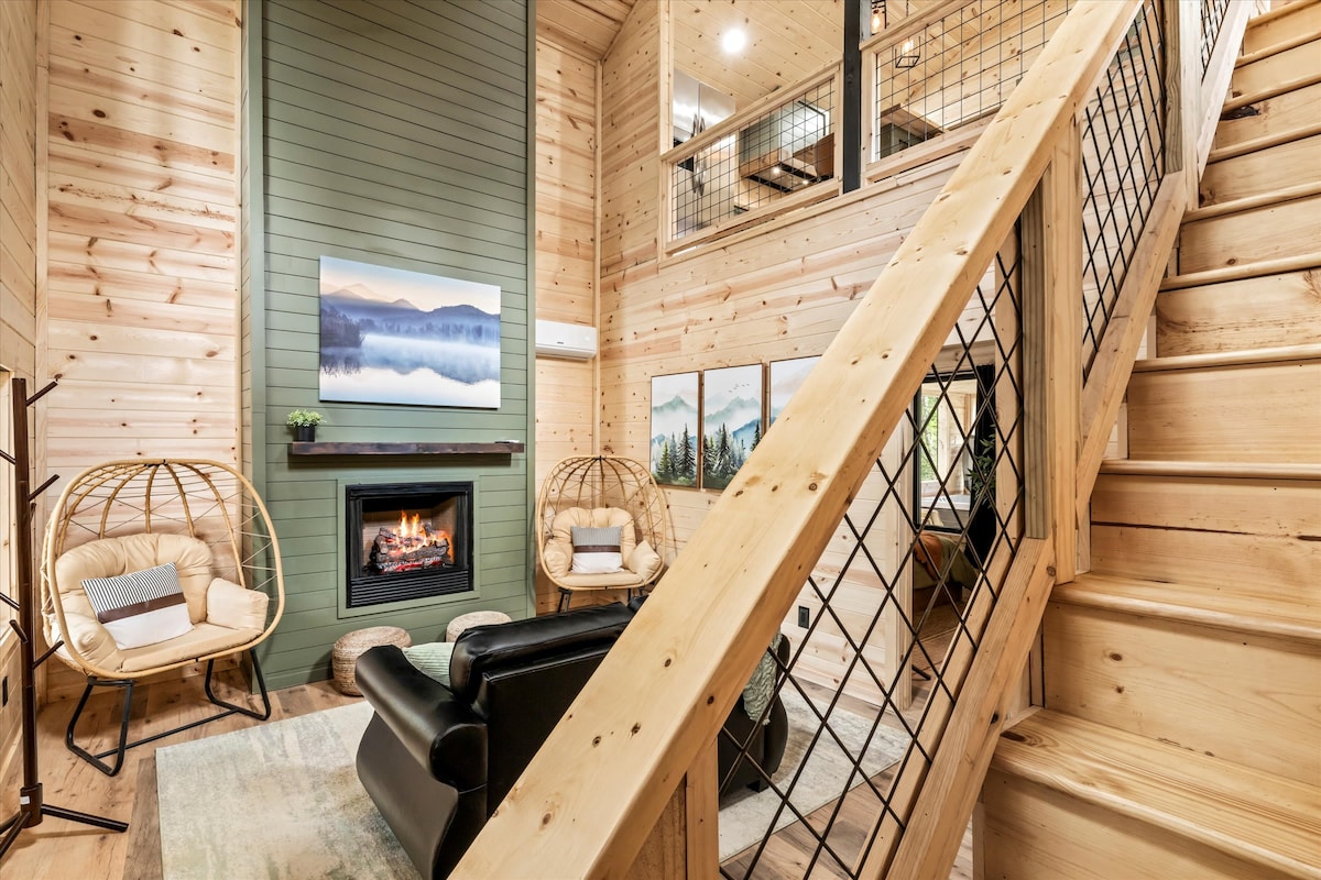 A welcoming living area features a black leather sofa and two cozy rattan chairs situated near a modern fireplace. A flat-screen TV is mounted above the fireplace, while light wood paneling and green accent walls contribute to the relaxed atmosphere. A staircase leads to the upper level.