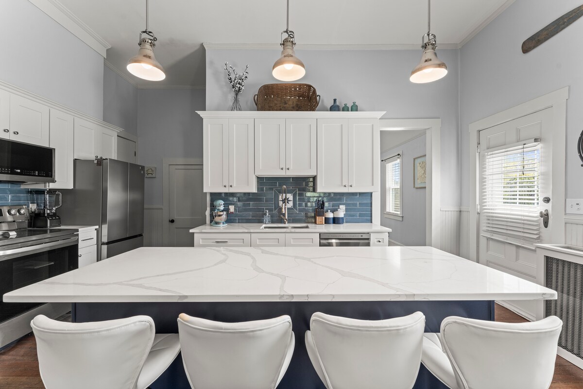 A modern kitchen features a large island with a white countertop, complemented by sleek white bar stools. Upper cabinets display a light color, while stainless steel appliances and a blue backsplash enhance the space. Warm light fixtures hang overhead, providing a welcoming ambiance.