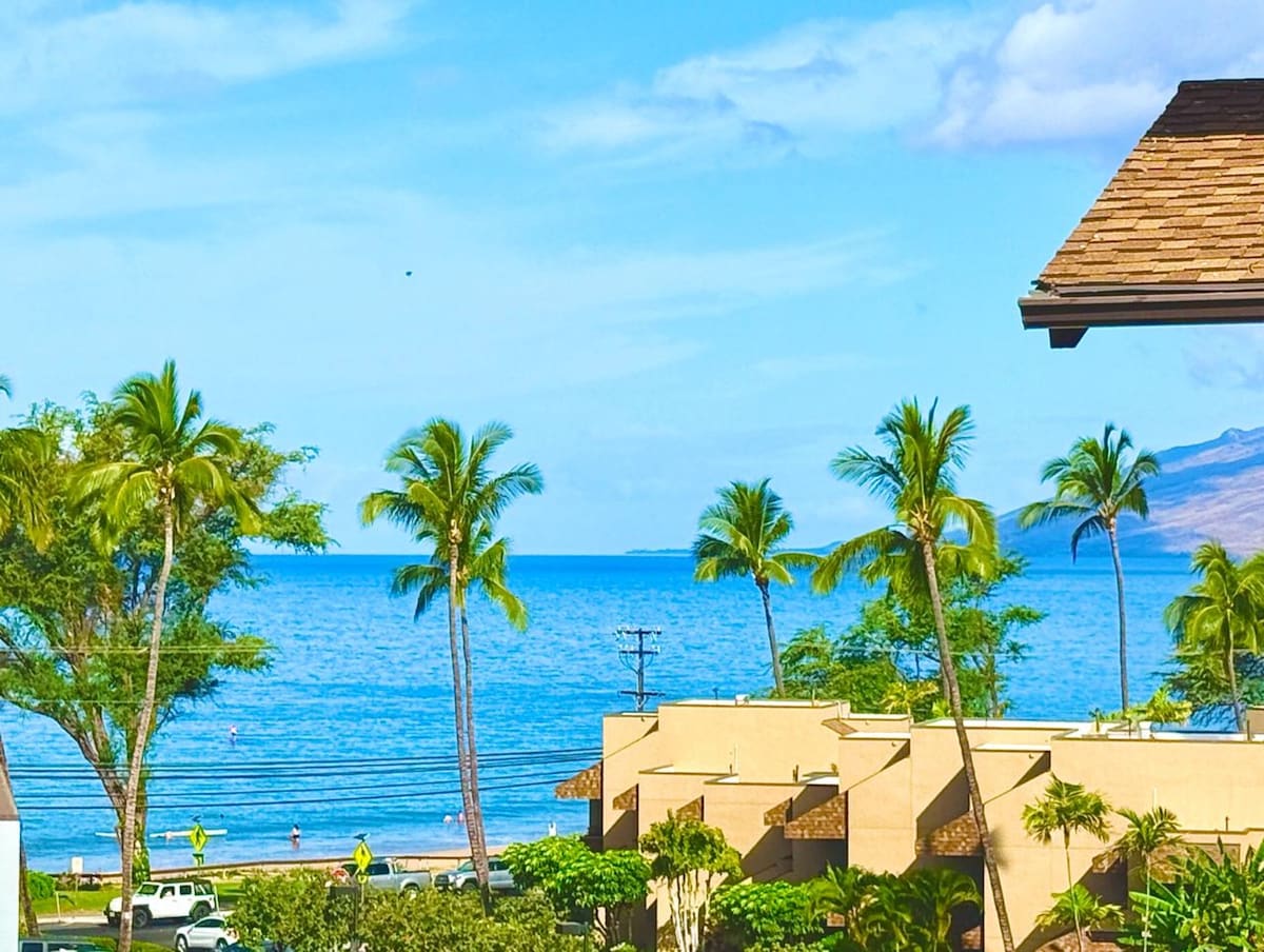 A panoramic view of the ocean and beach is framed by swaying palm trees. The clear blue water reflects a bright sky, while the shoreline is visible in the distance. The surrounding complex features lush greenery and modern architecture.