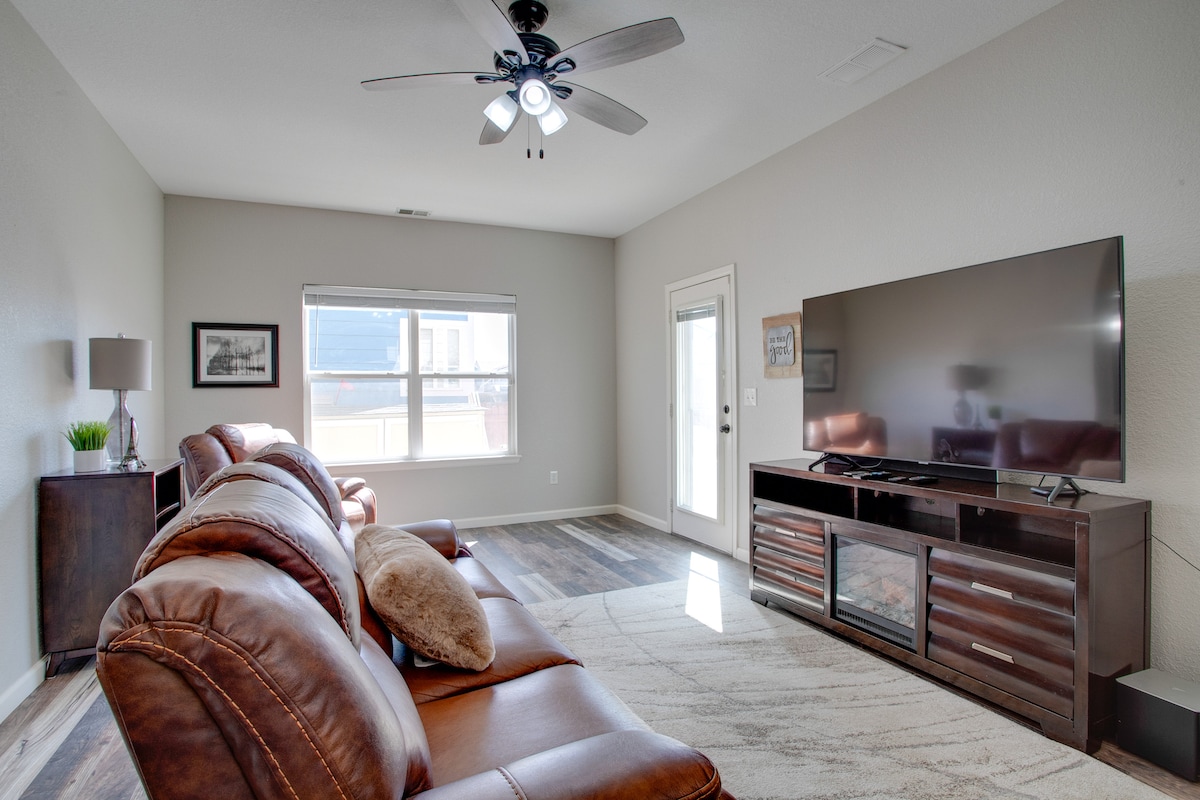 A comfortable living area is furnished with brown leather sofas and a large flat-screen TV mounted on a dark wood media console. Natural light enters through the window and patio door, highlighting the textured area rug on the floor.