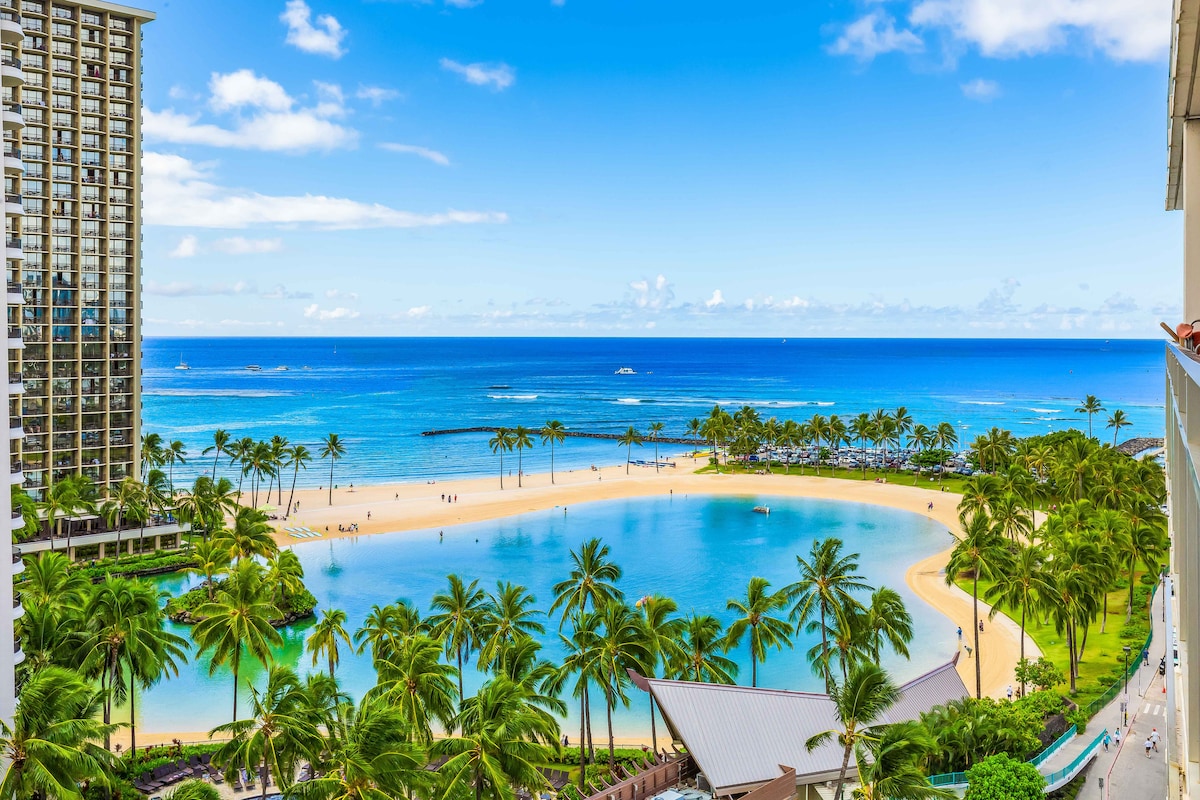 A sweeping view of a tranquil lagoon bordered by sandy beaches is captured, showcasing the vibrant blue ocean beyond. Palm trees line the shore, while beachgoers enjoy the sunlit water. The sky is a bright blue, dotted with soft white clouds, enhancing the scenic landscape.