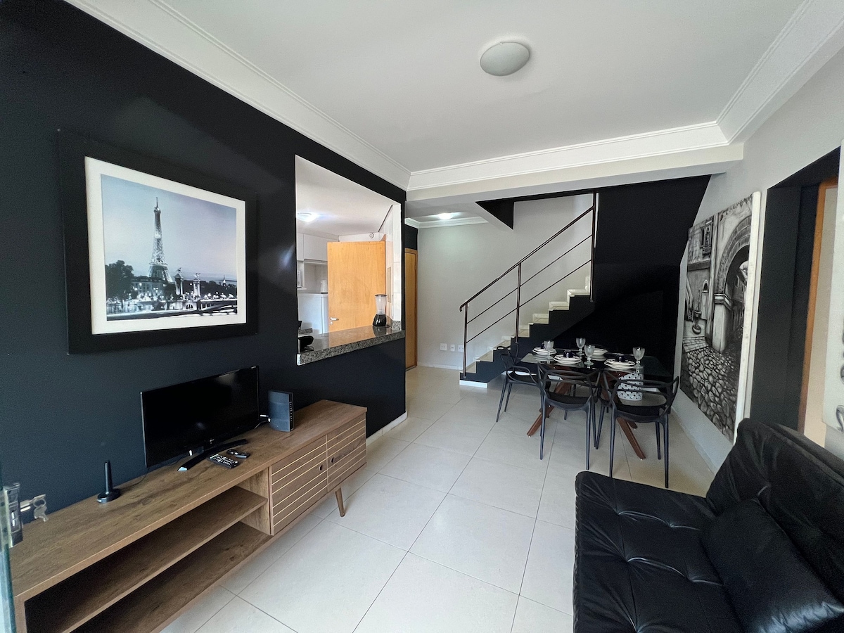 The living area features a sleek black wall and a contrasting light decor. A modern entertainment unit holds a television, while a dining table for four is set nearby. Natural light fills the space, and a staircase leads to the upper level.