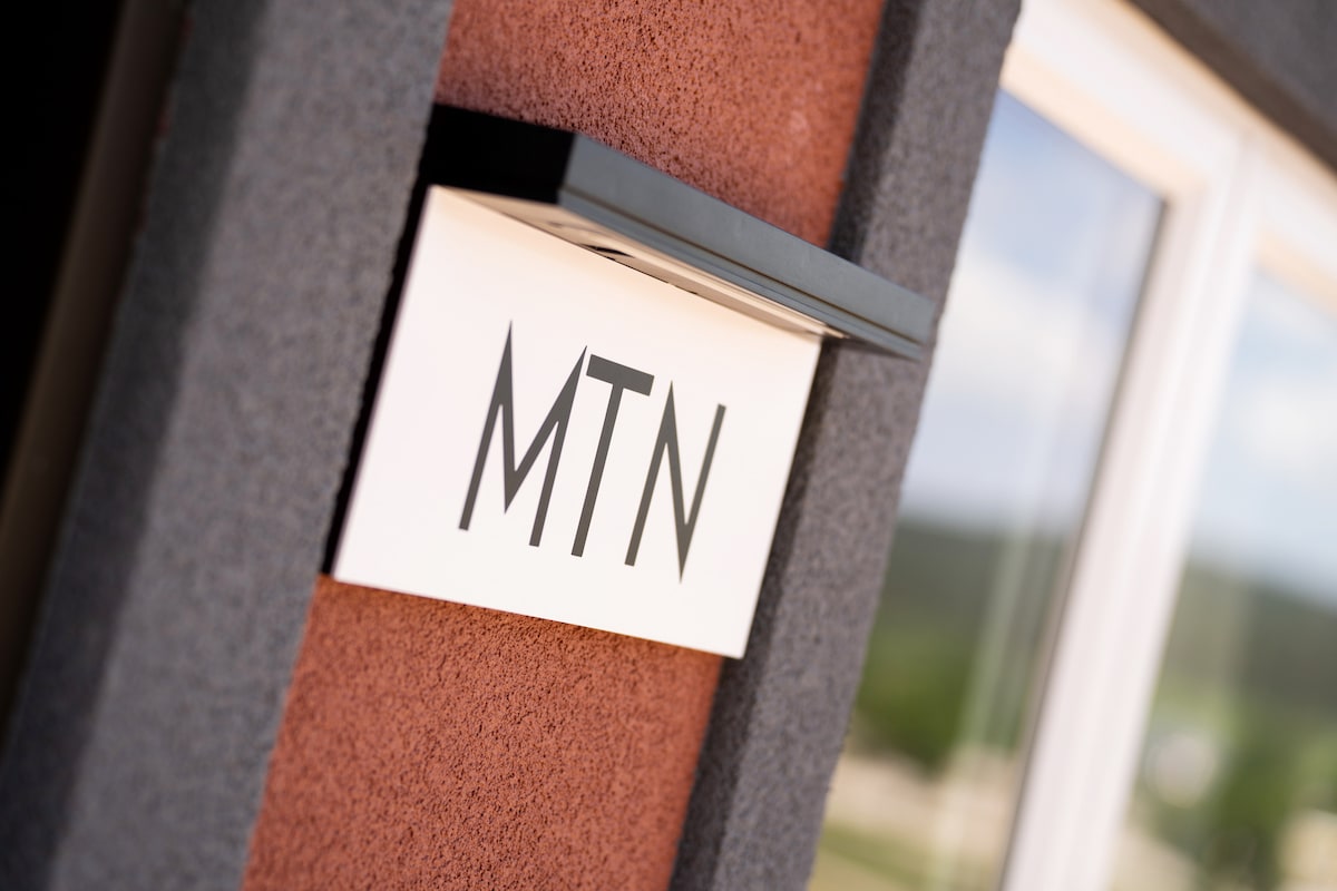 A close-up view of the exterior signage displaying 'MTN' in bold letters. The sign is mounted on a textured, colored wall, with a blurred backdrop of greenery and a large window visible.
