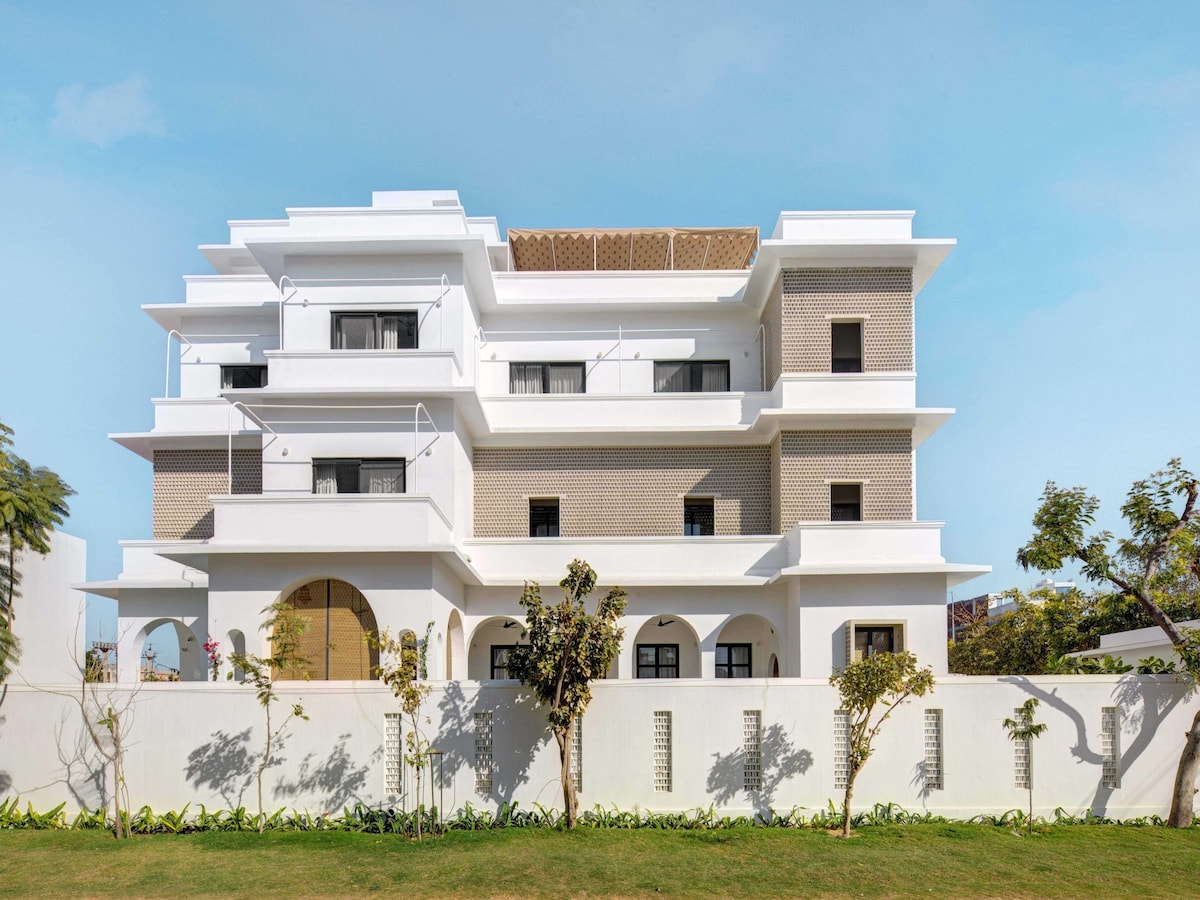 A modern, multi-level building with a combination of white and textured brick façades. Large windows and balconies are visible, set amidst a well-maintained garden area with greenery. The structure has a clean, contemporary design that integrates outdoor and indoor spaces.