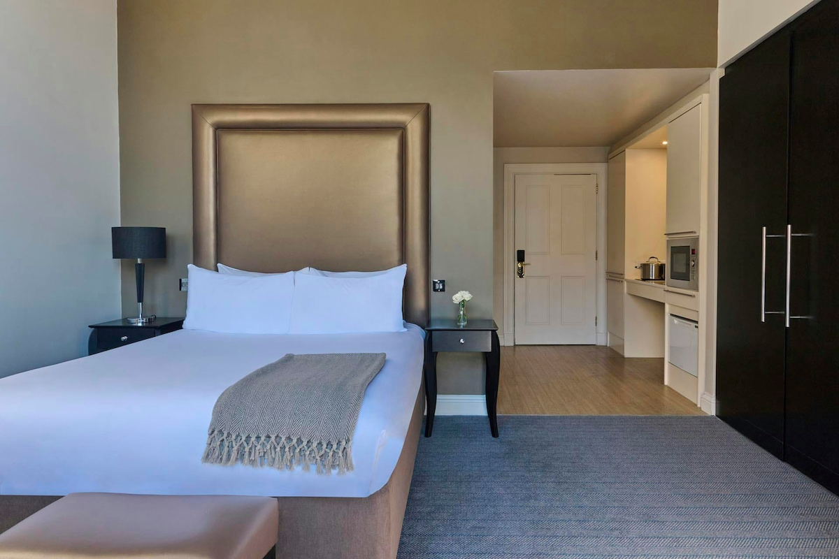A spacious studio contains a comfortable bed with a textured headboard, adorned with a neutral-toned throw blanket. A small table and lamp stand beside the bed. In the background, a well-equipped kitchenette is visible, featuring modern appliances and cabinetry.