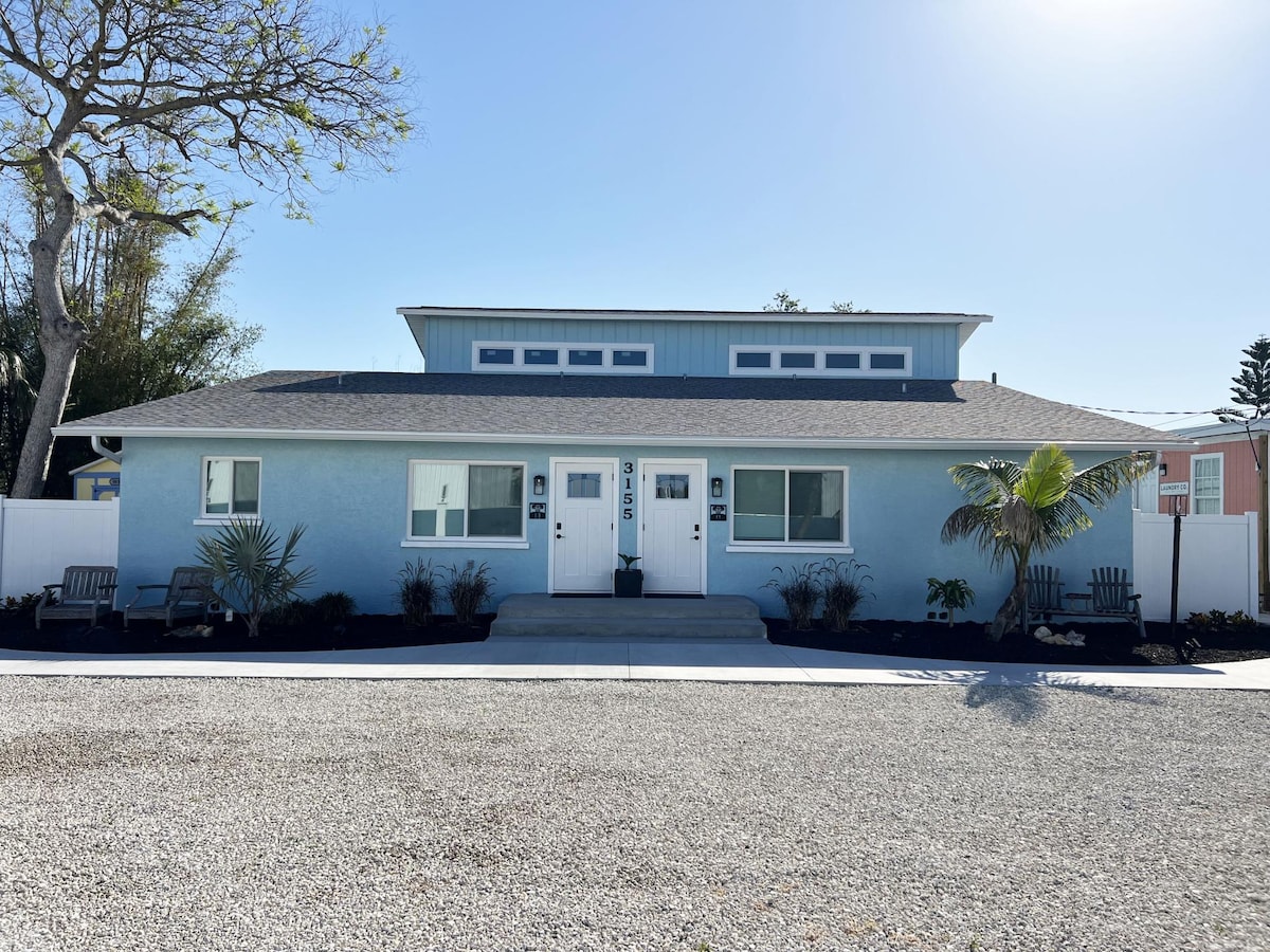 A newly remodeled retreat is visible, featuring a light blue exterior and a welcoming entryway. Two sets of double doors lead into the building, flanked by small seating areas. Lush greenery and decorative landscaping enhance the inviting facade under a clear blue sky.