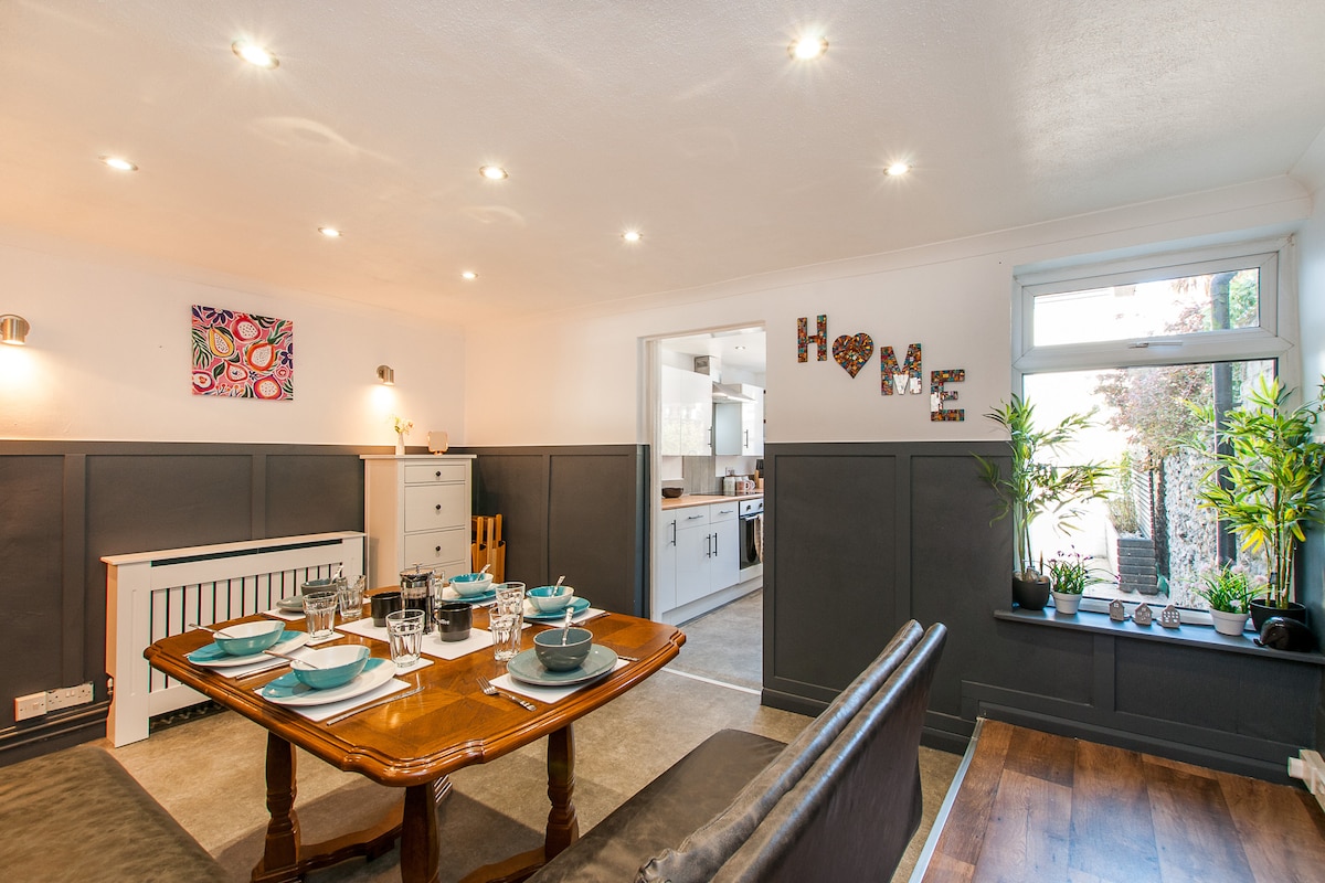 A dining area features a wooden table set for six, with blue dishware prominently displayed. The space is warmly lit, complemented by modern decor on the walls. A doorway leads to a modern kitchen, while a window offers natural light and a view of a small garden.