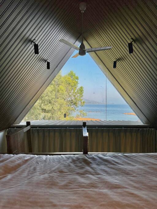 The loft bedroom features an angled ceiling and large triangular windows that frame views of Pawna Lake. Natural light spills into the room, highlighting the soft bed linens. Lush greenery is visible outside, complementing the tranquil water scene beyond.