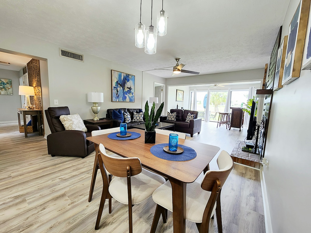A spacious open-concept living area features comfortable seating arrangements, including a dark leather couch and armchair. A dining table set for four is visible, alongside an elegant light fixture and tasteful artwork. Large windows allow natural light to brighten the space, enhancing its inviting atmosphere.