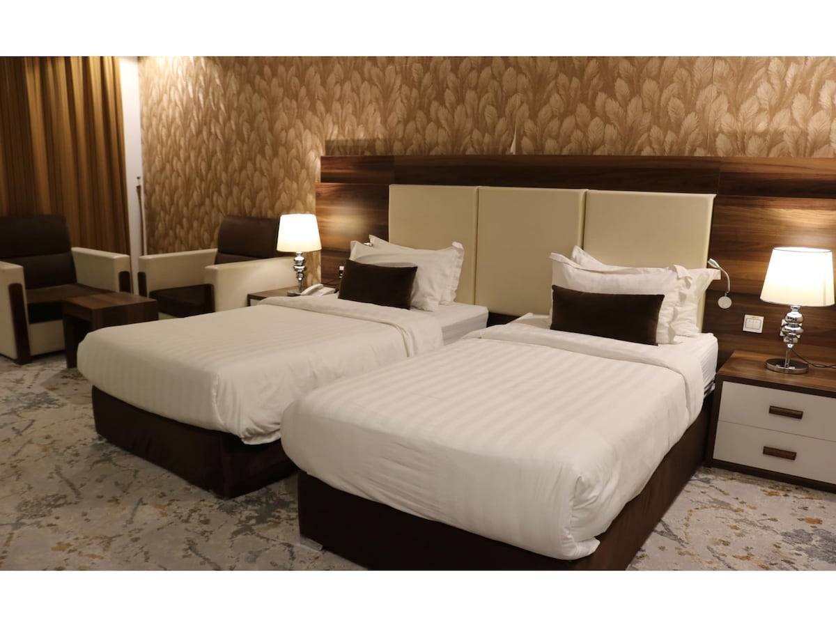 Two spacious twin beds are positioned in a well-lit room with soothing wall decor. Each bed features a plush headboard and is accompanied by bedside lamps. A cozy seating area is present, enhancing the inviting atmosphere of the room.
