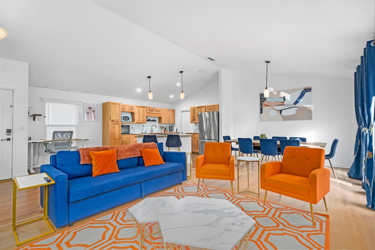 An open living space showcases a vibrant blue sofa accompanied by orange accent chairs. A marble coffee table sits in the center. The adjoining kitchen features wooden cabinetry and stainless steel appliances, while a dining area is furnished with a table for six and additional seating.