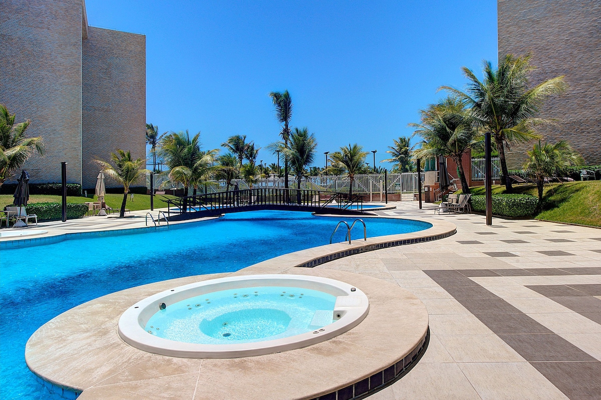 A large swimming pool is surrounded by well-maintained grass and palm trees, offering a serene setting. A whirlpool is positioned nearby, both providing a relaxing experience under a clear blue sky. Lounge chairs are arranged around the pool area for guest comfort.