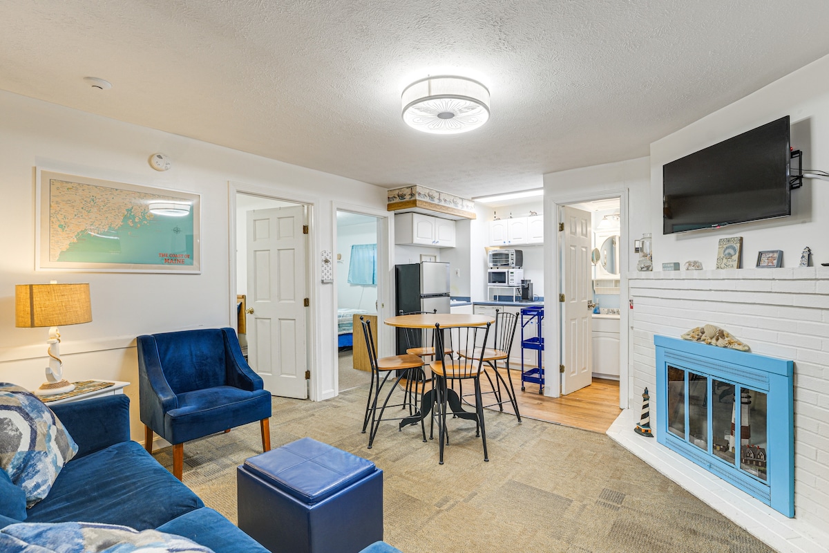 A cozy living area is showcased, featuring a blue armchair and matching ottoman alongside a sectional sofa. A round dining table holds four black chairs. A flat-screen TV is mounted above a decorative fireplace with a pastel blue mantel, creating a welcoming space.