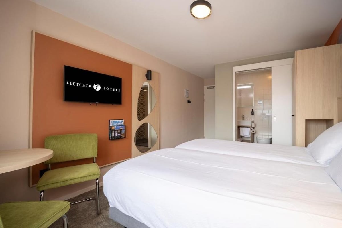 A modern hotel room is presented with two spacious beds, a flat-screen TV mounted on the wall, and a round table paired with a green chair. A partition reveals a private bathroom, and natural light enters through a nearby window.
