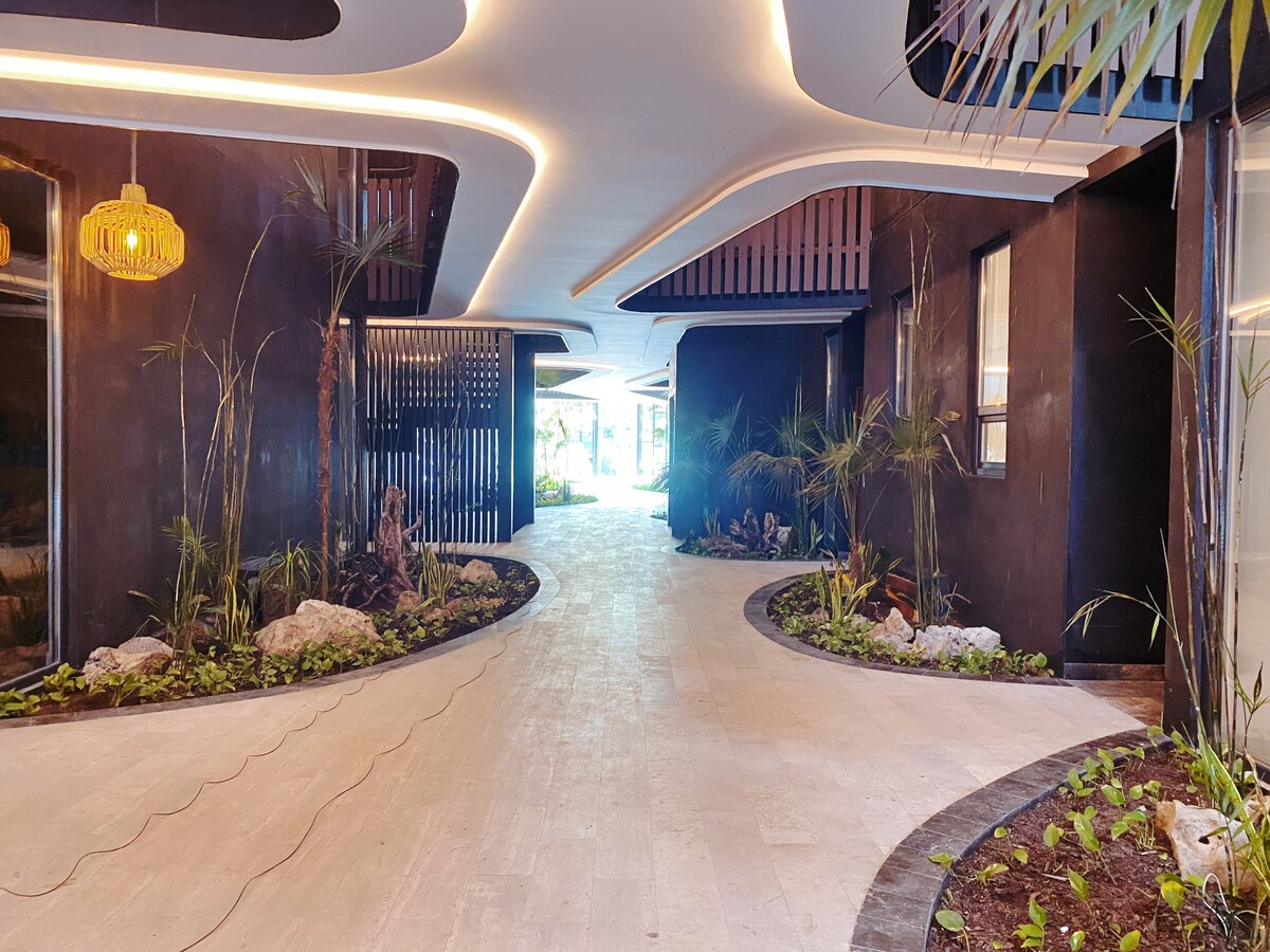 A modern entryway is presented, featuring a smooth pathway lined with lush greenery and decorative stones. Ambient lighting is provided by woven pendant lights. The stylish architecture encompasses elegant curves and contemporary design elements, creating a welcoming transition from the outdoors to the interior.